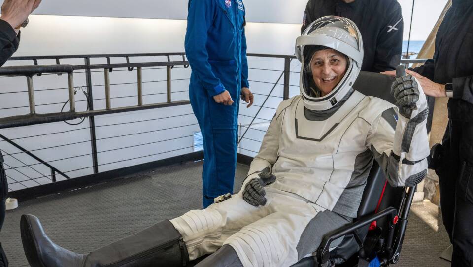 A woman in a space suit smiles as she sits on a stretcher.