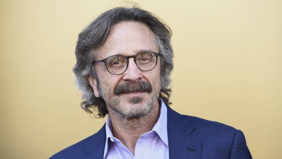 Marc Maron, wearing a button-up shirt and jacket, smiles slightly as he looks ahead.