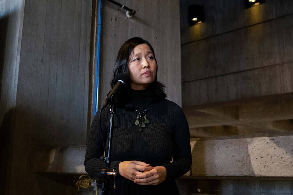 A woman in a black sweater dress holds her hands in front of her and looks off with a serious expression.