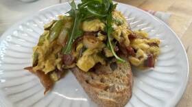 White plate topped with toasted sliced bread with egg and prosciutto with arugula on top