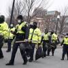 a group of state troopers walks in boston, wearing warm black clothes like hats and layers under bright yellow vests, carrying sticks, helmets and other gear while wearing masks
