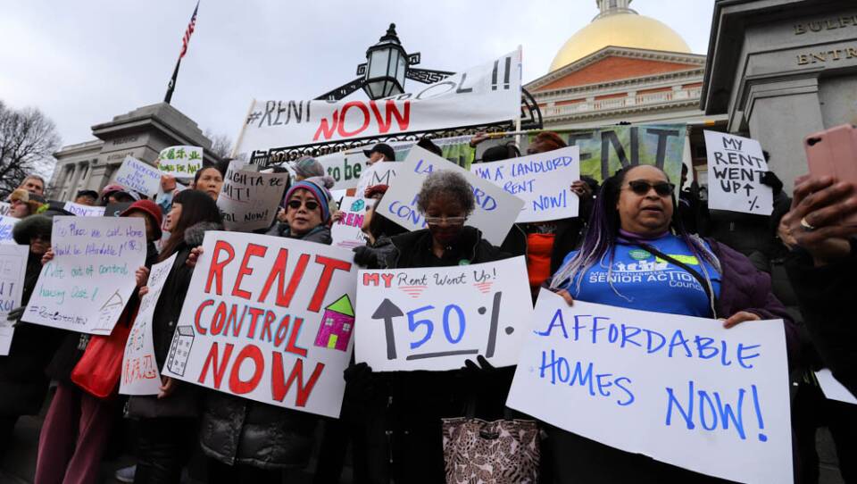 A group of people hold signs in favor of rent control and affordable housing outside of the gold-domed Massachusetts State House.