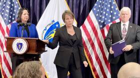 A woman with short brown hair, wearing a black suit, stands with her hand on a wooden lectern. Flags and two other people are behind her.