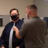 In a doctor's office, a person holds up a stethoscope to a person's chest. Both are wearing masks.