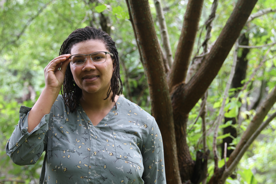 A woman, half-smiling, stands amid greenery and adjusts her glasses on an overcast day