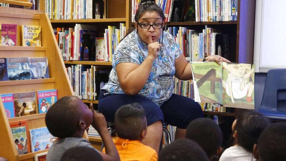 A women sits in a chair in a library full of books, her finger to her mouth, reading aloud from an illustrated book to a diverse group of children, who are sitting on the floor.