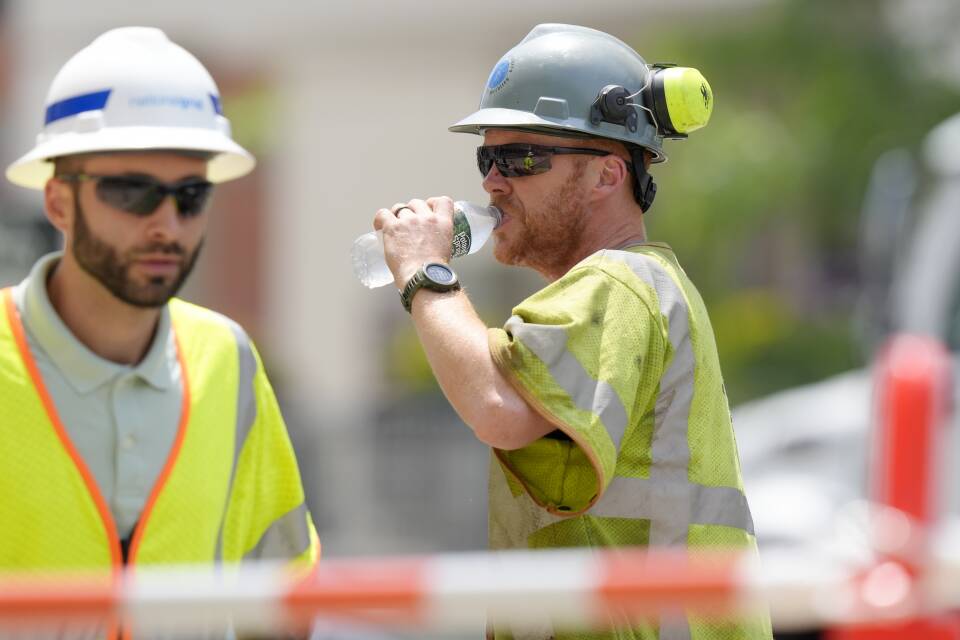 A person wearing a high-visibility shirt and helmet drinks from a bottle of water as the sun beats down.