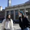 Two young adults smile as they talk while walking across Boylston Street. They have masks lowered around their chins. A handful of other pedestrians are also crossing the street, some in masks.