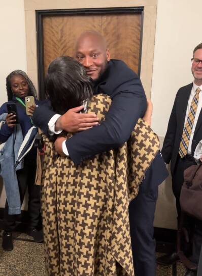 A black man in a suit hugs a black woman wearing a beige plaid wrap.