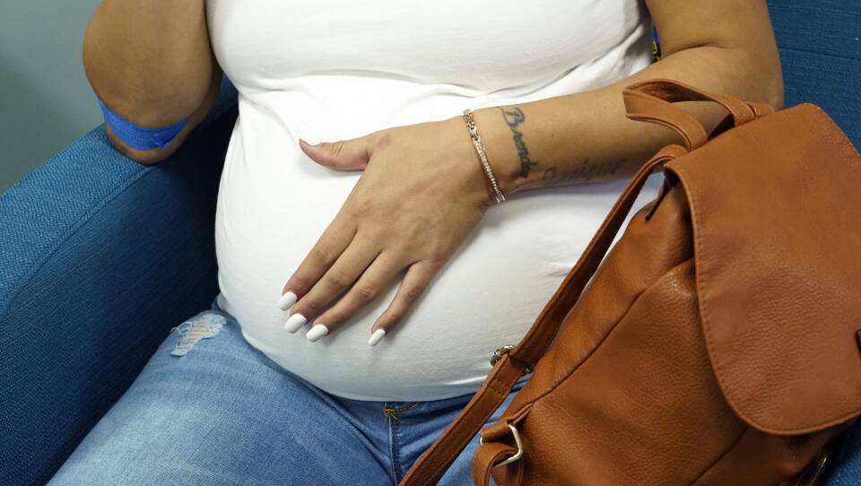 A pregnant woman rests one hand on her belly as she sits on a chair in a waiting room.
