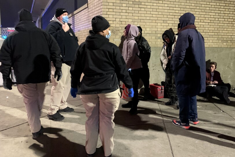 Three people in khaki pants and black jackets stand on the sidewalk at night as they speak with five other people standing and sitting around the edge of a building.