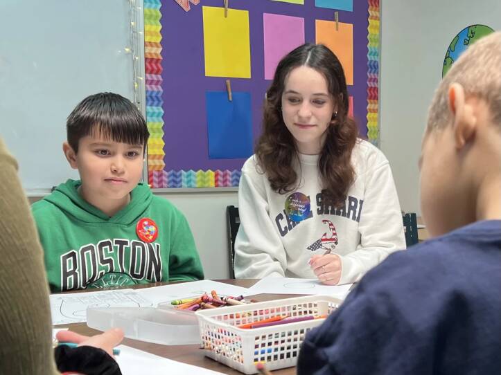 A young boy in a green Boston Celtics sweatshirt sits with a box of crayons and colored pencils in front of him. A teenage woman wears a white sweatshirt and sits next to him smiling with no teeth.