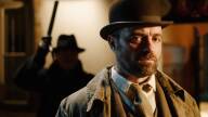 A man in a bowler hat looks into the camera. Behind him, in silhouette, is another man, appearing to be ready to attack.