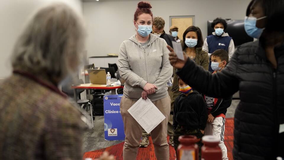 A woman wearing a blue surgical mask stands at the front of a short line of people, including two children and adults of varying ages.