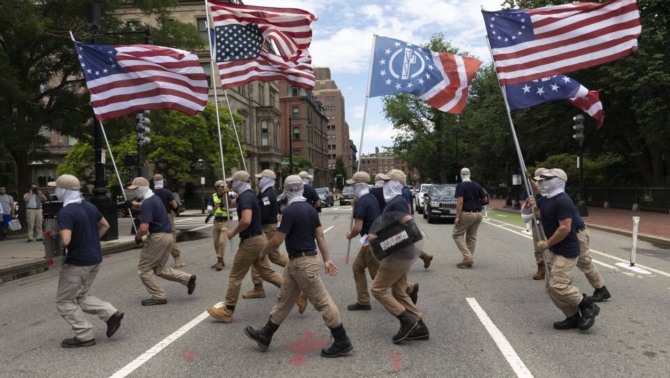 A dozen men are shown with matching face coverings, navy shirts, khaki pants and khaki baseball caps. They're waving various American flags and the Patriot Front insignia.