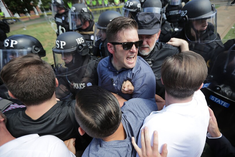 Police in riot gear push in on a small group of men, one at the center wearing sunglasses and a button-down shirt, yelling