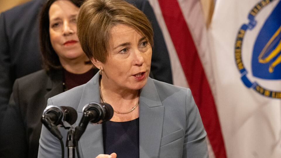 A woman in a blazer speaks intently into a microphone, with flags and assorted other officials in the background.