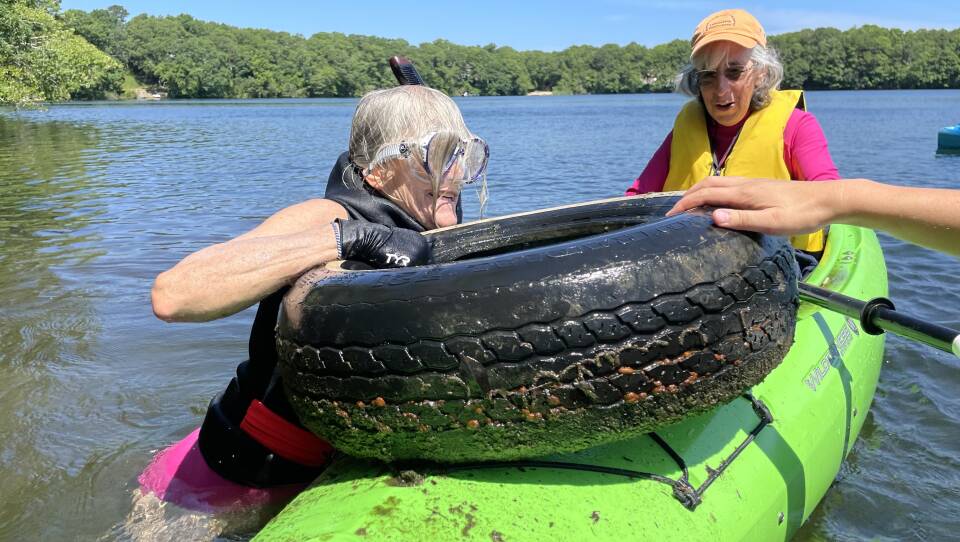 An older woman in a swimsuit and goggles lifts a tire onto the front of a green kayak that another older woman is paddling.