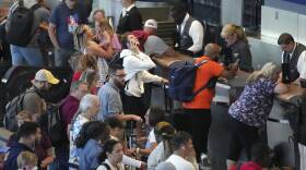 A crowd of people with luggage look upset and frustrated while waiting at an airport service counter.