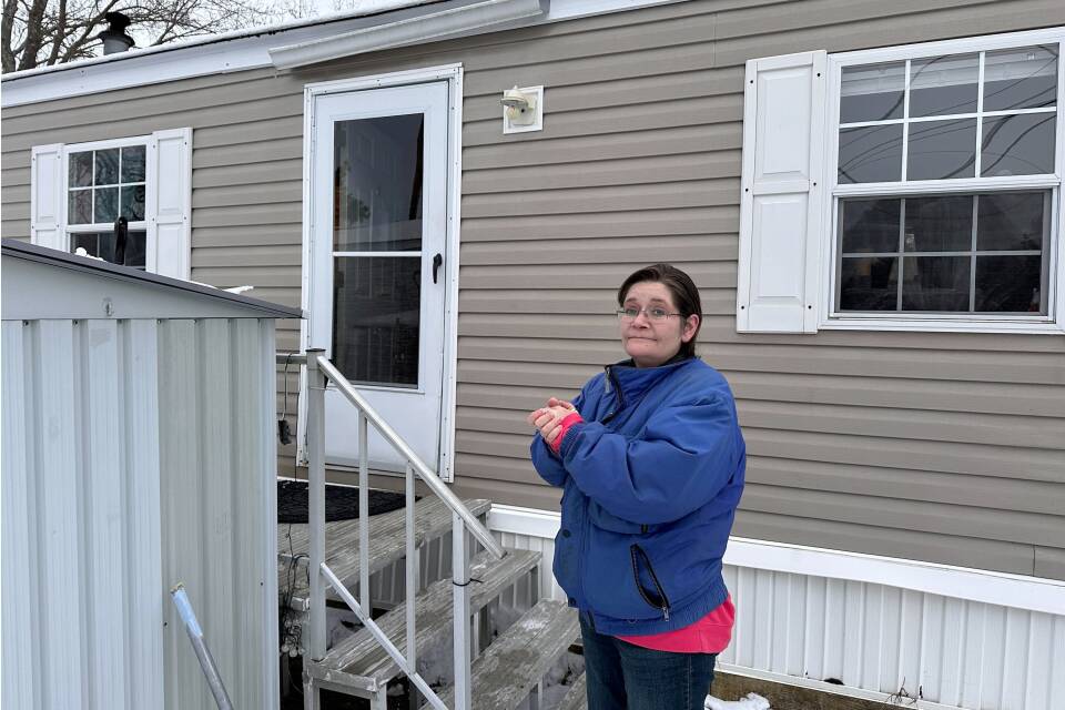 A woman stands by the stairs outside a mobile home. She looks sad and is holding her hands together.