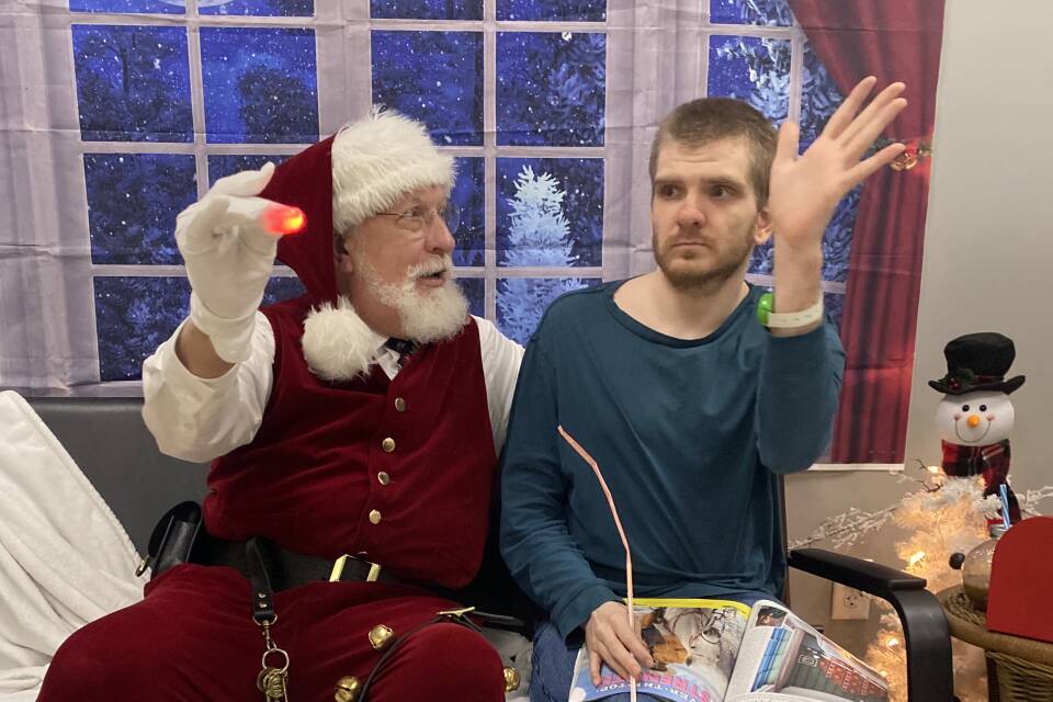 A man waves his hand as he sits on a couch next to Santa Claus.