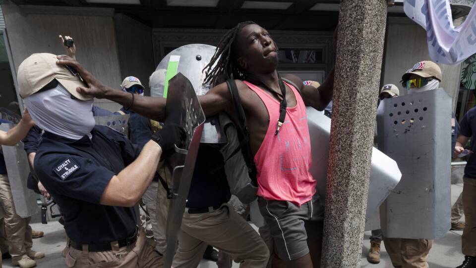A group of masked men in matching clothes and face coverings use metal shields to push a Black man up against a post on the sidewalk.  The men are members of the Patriot Front, a neo-Nazi group formed after the Unite The Right violent rally in 2017.