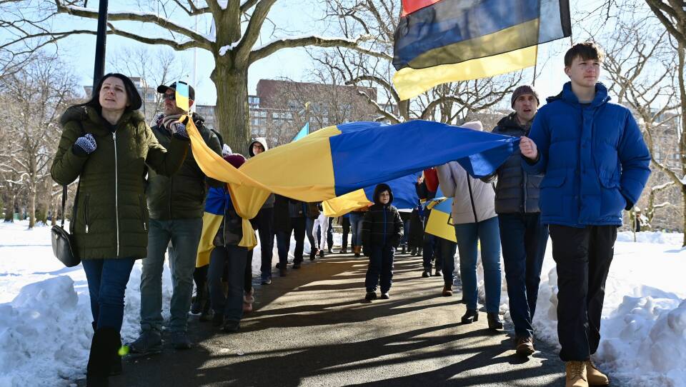 Two rows of people walk on the sides of a cleared path, holding the blue and yellow Ukrainian flag between them, while a young boy walks between them under the flags