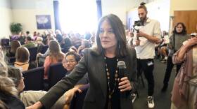 Marianne Williamson holds a microphone as she walks around the back of a conference room full of people.