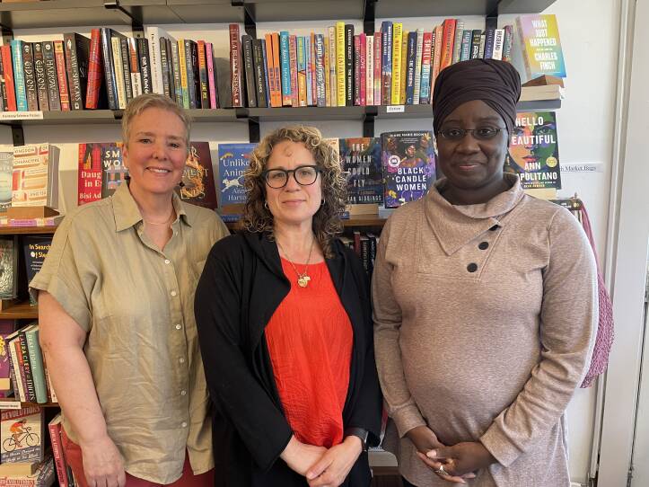 Three women stand against a bookcase in a bookstore. They are all smiling