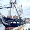 The USS Constitution, a wooden 18th Century warship with three tall masts, sits with her sails down, tied alongside a pier.