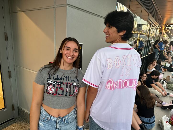 A young woman smiles with teeth and a young man smiles with teeth, the woman wears a T-shirt that says "I (don't) hate Boston" and the young man wears a white jersey that says "Boston is Renough"