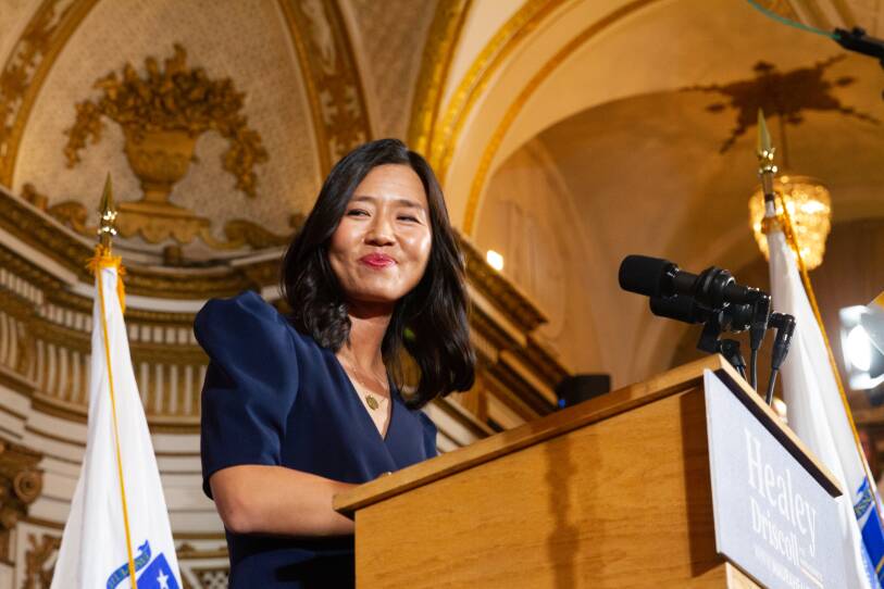 A woman in a dark blue dress smiles from behind a podium in a fancy ballroom as she looks to the side.