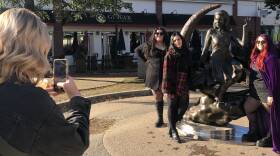 Three women, in a black dress, red and green plad coat and skirt, and a long purple cape, pose in front of a statue of a woman sitting on a half-moon.