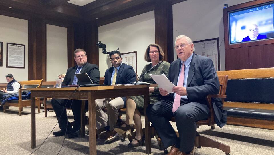 Three men and a woman sit in front of microphones in a hearing room, with several benches behind them.