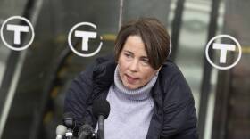 A woman in a turtleneck sweater and coat speaks into a stand full of microphones outside of an MBTA station.