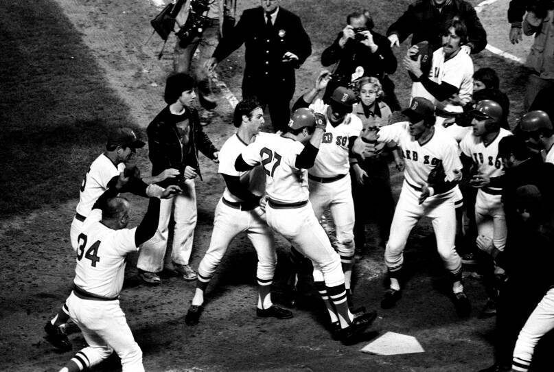 A baseball player jumps with both feet onto home plate as a crowd of teammates cheer around him.