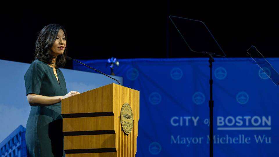 A woman stands behind a podium against a backdrop that has the seal of the City of Boston and a picture of Boston City Hall.