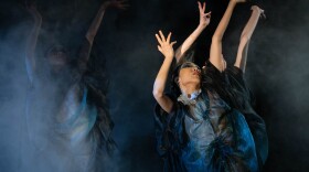 Three dancers are wearing diaphanous blue and white gauze dresses. Their arms are outstretched over their heads as they look upwards. The stage is misty with fog and blue light