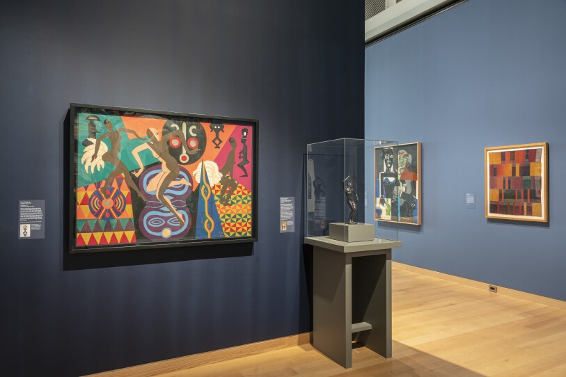 This is a photograph of a museum installation. Hanging against a dark blue wall is a collage with geometric shapes: triangles, circles, and dancing figures. The colors are vibrant with yellows, reds, blues and purples. To the right of the paining is a small Benga Dance figure made of bronze. It's displayed in a glass case.