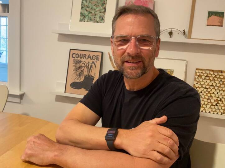A man sits at his kitchen table, arms crossed and wearing a smart watch. Framed artwork is displayed on the wall behind him showing abstract plants and also one illustration with the word "courage."