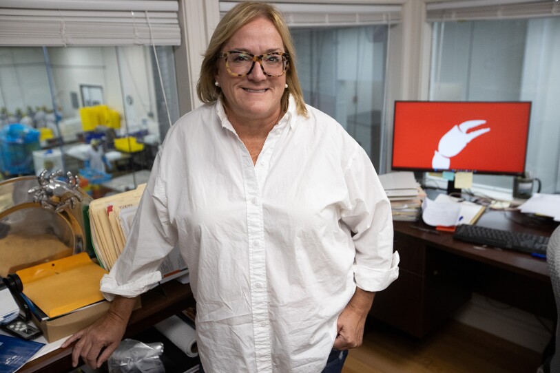 A woman in a white buttondown smiles for the camera. She's in an office, with a factory visible through the background through her window.