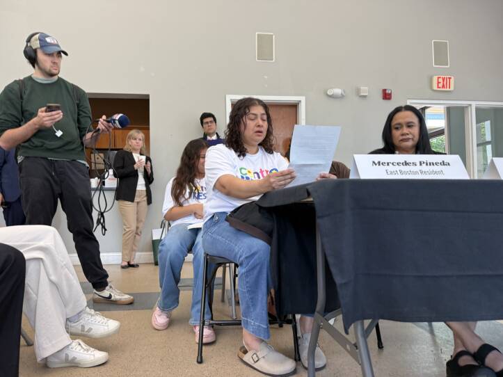 A woman sits at a table and looks at a paper as she speaks.