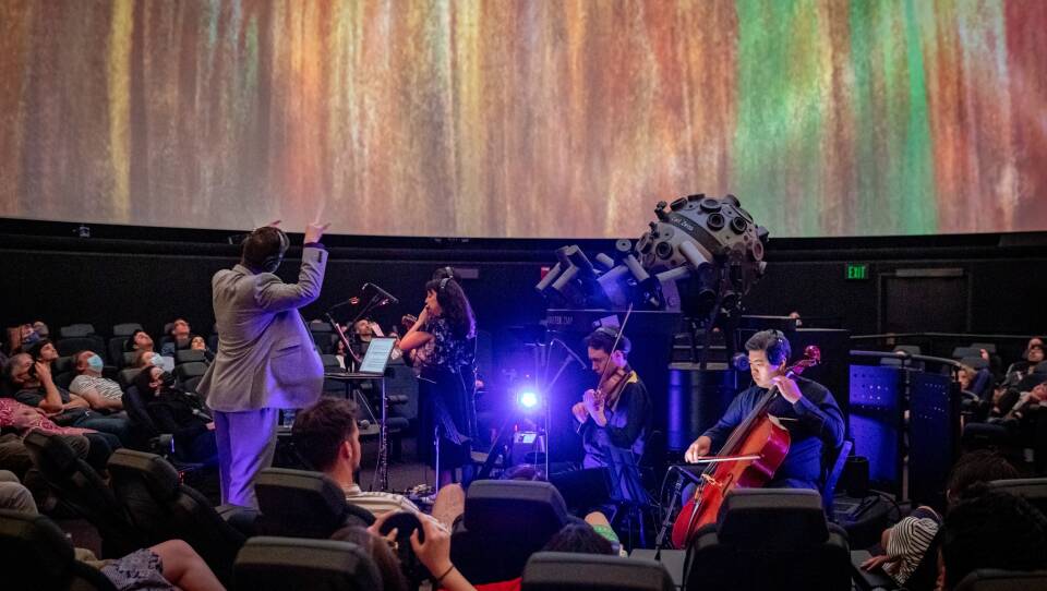 A conductor leads musicians in the center of a small planetarium, surrounded by audience members whose eyes are fixed on the ceiling where bright animated colors fill the screen