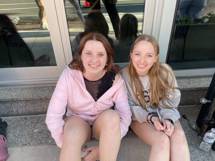 Two young women are sitting outside on a sidewalk. They are smiling as they wait to get into a concert.