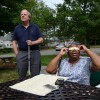 In a suburban yard, a man and a woman both hold white canes. On a table, are a smartphone and a booklet of raised-line, tactile images of a solar eclipse.