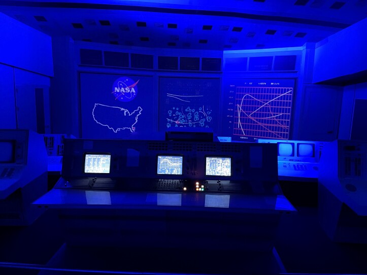 This photograph is of a recreation of a NASA control room with an original switchboard. The room is bathed in dark blue light. In the foreground is a console with three monitors and control panels. In the background is an illuminated display of charts, a map of the United States and the NASA logo