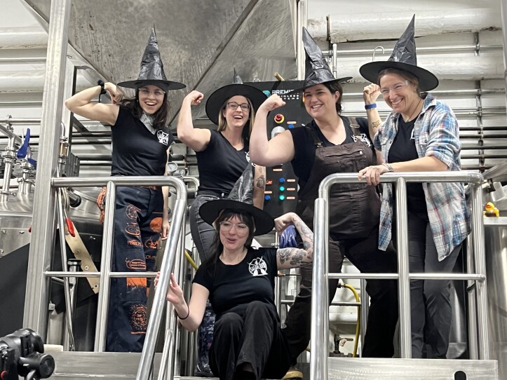 Women wear black shirts and witch hats and flex their muscles.