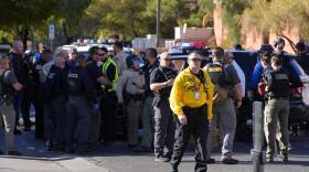 More than two dozen law enforcement officials stand on a campus on a sunny day. Police cars are in the background.