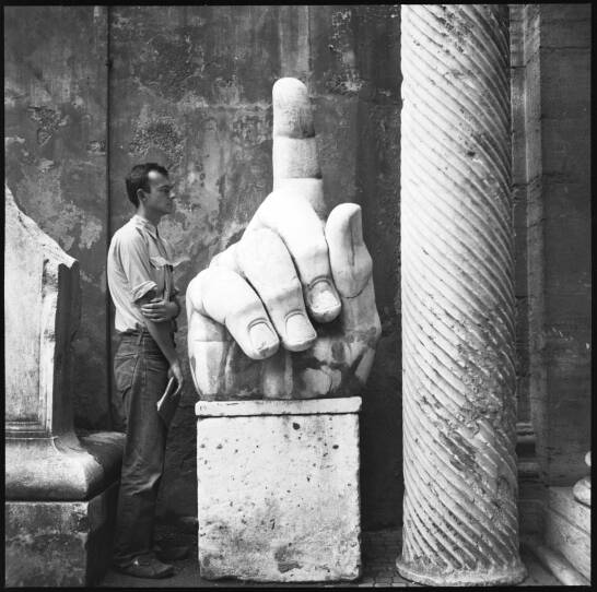 A black and white photograph shows artist Cy Twombly looking at a column and giant sculpted hand