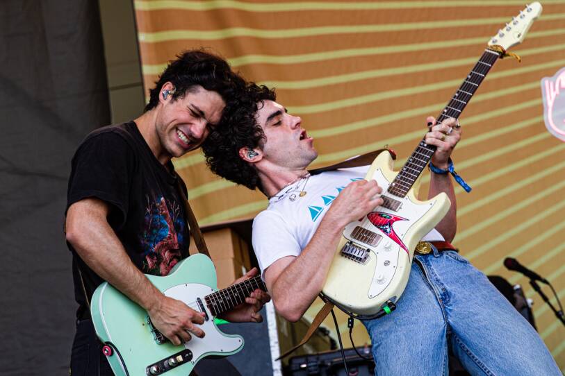 This is a photograph of singer and musician Del Water Gap performing onstage with a backup guitarist at the Newport Folk Festival. As Del Water Gap plays his electric guitar, he leans back with his eyes closed. The back of his head is touching the forehead of the backup guitarist who is standing directly behind Del Water Gap as he plays his electric guitar.